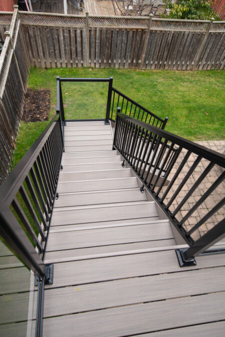 A view looking down a set of modern outdoor stairs with gray composite decking and black metal railings, leading from a deck to a grassy backyard enclosed by a wooden fence.