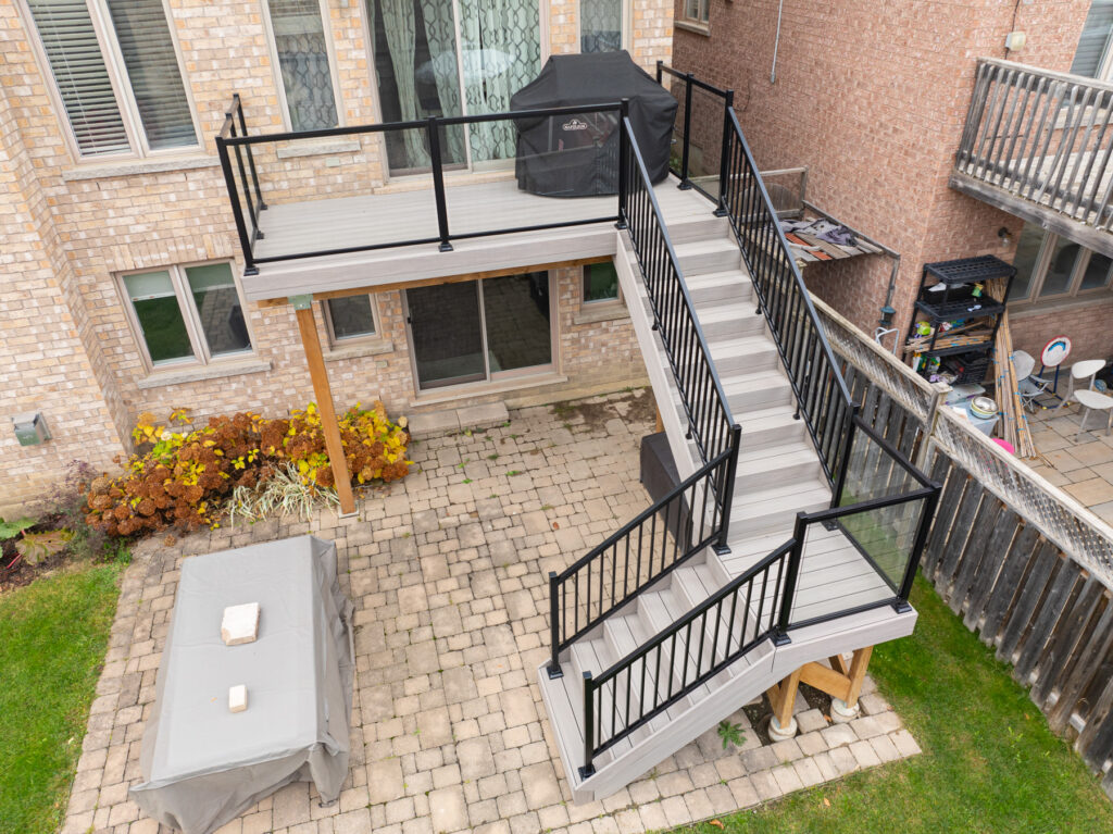 Aerial view of a two-story backyard deck with black railings, stairs leading down to a patio area with a covered table, paver stones, grill, and plants along a brick house.