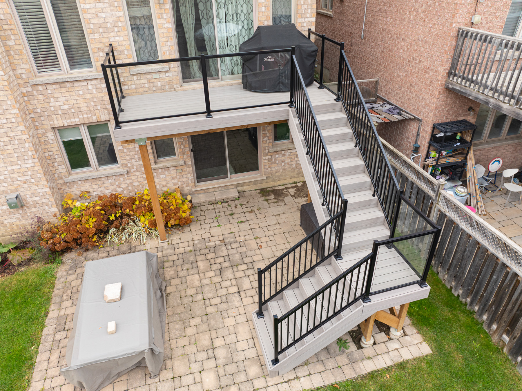 Aerial view of a two-story backyard deck with black railings, stairs leading down to a patio area with a covered table, paver stones, grill, and plants along a brick house.