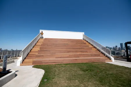 Large wooden steps lead up to a rooftop platform with metal railings; a person stands at the top under a clear blue sky.