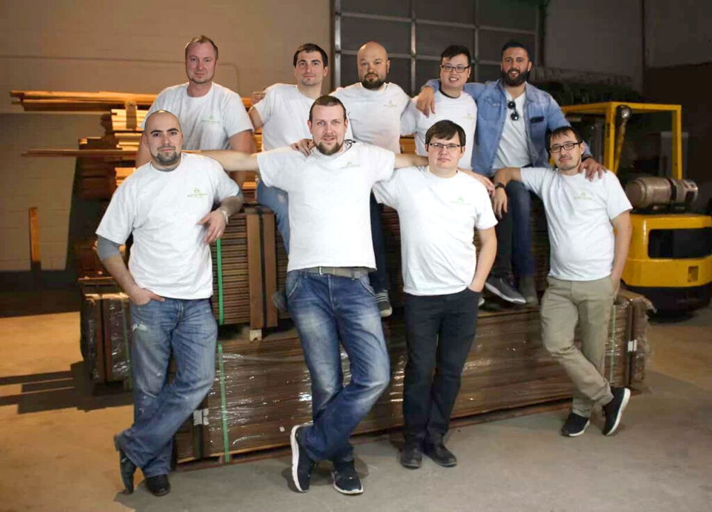 Nine men from the Delta Decks team, dressed in white t-shirts and jeans, pose together in a warehouse, standing and sitting on stacked wooden planks. A yellow forklift is visible in the background.