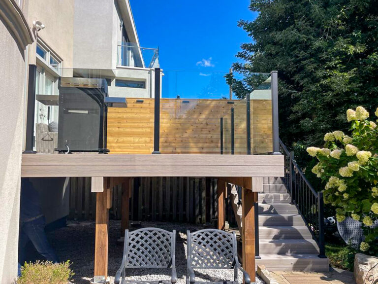 A modern raised deck with Century Scenic glass and aluminum picket railings, plus a wooden privacy screen, features stairs descending to a shaded area with two lounge chairs. Flowering bushes and trees surround the patio on a sunny day.