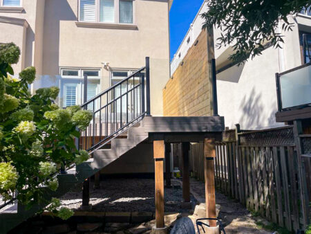 A modern raised deck built with Fiberon Composite Decking features black railings and stairs, attached to the back of a beige house and overlooking a small gravel yard with plants, trees, and a wooden fence.