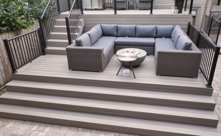 A modern outdoor patio with Fiberon Composite Decking features a sunken seating area where gray cushioned wicker sofas form an L-shape around a round coffee table, surrounded by stairs and sleek black railings.
