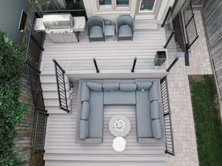 Aerial view of a modern backyard deck with Fiberon Cabana boards, a gray L-shaped sofa, round white table, two armchairs, barbecue grill, and black railings—overlooking a paved patio and lush green grass.