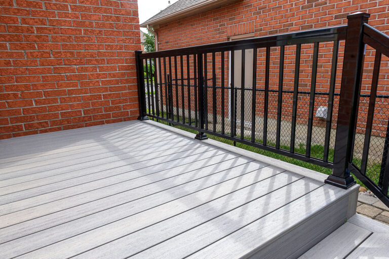 A modern deck with light gray composite flooring and a black metal railing, attached to a red brick house. In the background, there is a chain-link fence and another brick building.