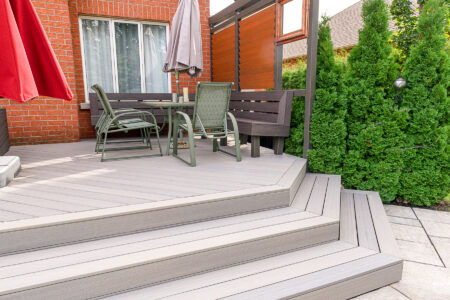 A modern backyard patio with gray composite decking, green chairs, a bench, a table, large red and gray umbrellas, and privacy fencing, surrounded by green hedges and a brick house.