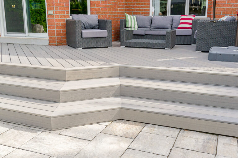 A modern outdoor patio featuring light gray composite deck steps, gray wicker furniture with gray cushions, and colorful accent pillows, set against a red brick house and stone paver patio.
