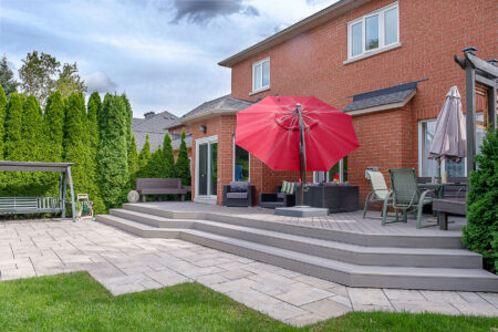A backyard patio with a raised deck featuring outdoor furniture, a red umbrella, a dining table with chairs, a swing bench, and green shrubs bordering a brick house under a partly cloudy sky.