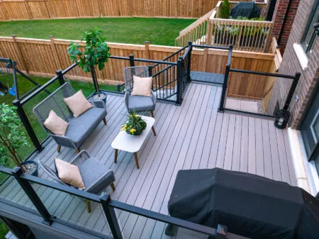Aerial view of a Fiberon Chai deck with Fortress-Railings1 showcases gray flooring. Features include a seating area with gray chairs and beige cushions, a white coffee table with plants, another decorative plant, and a covered grill. Its all beautifully enclosed by a wooden fence and lush lawn.