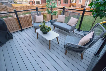 Side-by-side images of the Fiberon modern outdoor staircase with gray steps and black railings leading to a raised patio. The patio is bordered by glass panels and decorated with potted plants. The setting includes brick walls and a paved walkway.