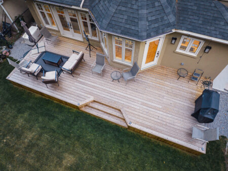 Aerial view of a natural wood decking backyard deck attached to a house, featuring outdoor furniture including sofas, chairs, tables, and a grill, with steps leading down to a grassy lawn.