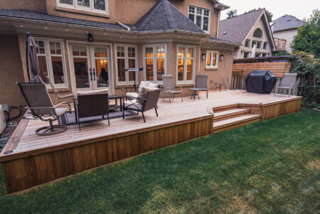 A wooden backyard deck with patio furniture, including chairs and tables, bordered by green grass. String lights hang above, and a grill is on the right side near a wooden fence. The deck is attached to a beige house with large windows.