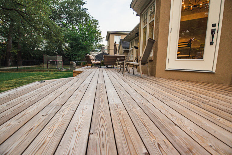 A spacious Lunawood Decking attached to a house, featuring several chairs and a table for outdoor seating. The deck overlooks a grassy backyard dotted with trees and fenced for privacy.