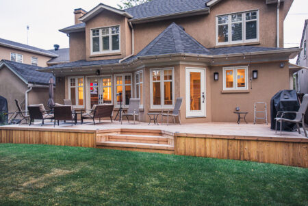 A spacious Lunawood backyard deck with outdoor chairs, tables, and a grill, attached to a two-story tan house with many windows and a well-kept lawn.