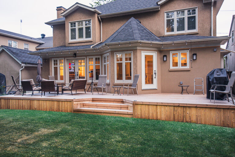 A spacious Lunawood backyard deck with outdoor chairs, tables, and a grill, attached to a two-story tan house with many windows and a well-kept lawn.