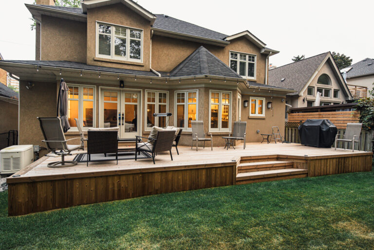 A large house with a tan exterior, many windows, and a spacious backyard deck made of Lunawood Natural Wood decking, furnished with patio chairs, a table, an umbrella, and a grill, overlooking a green lawn.