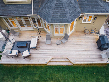 Aerial view of a Lunawood wooden backyard deck attached to a house, featuring outdoor seating, a table, two round tables, a barbecue grill, and lush green grass surrounding the deck.
