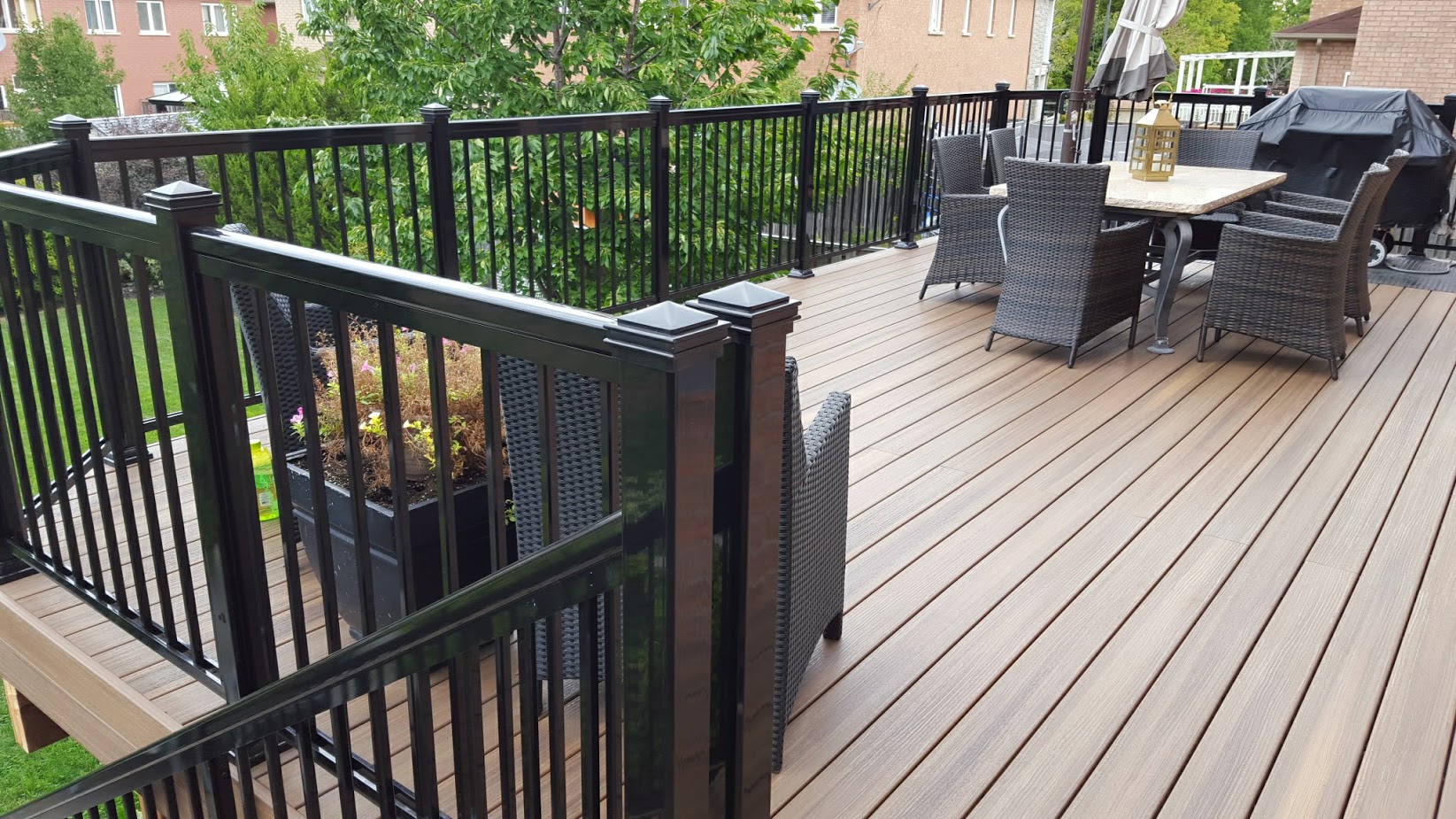 A spacious wooden deck with black railings, outdoor dining furniture, a potted plant, and a grill, overlooking a green yard and brick houses.