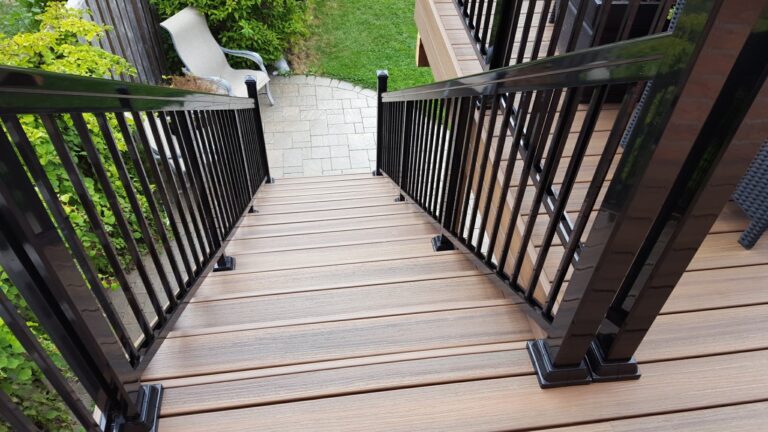 A view looking down a set of brown composite deck stairs with black metal railings, leading to a paved patio area and green lawn with plants and a lounge chair.