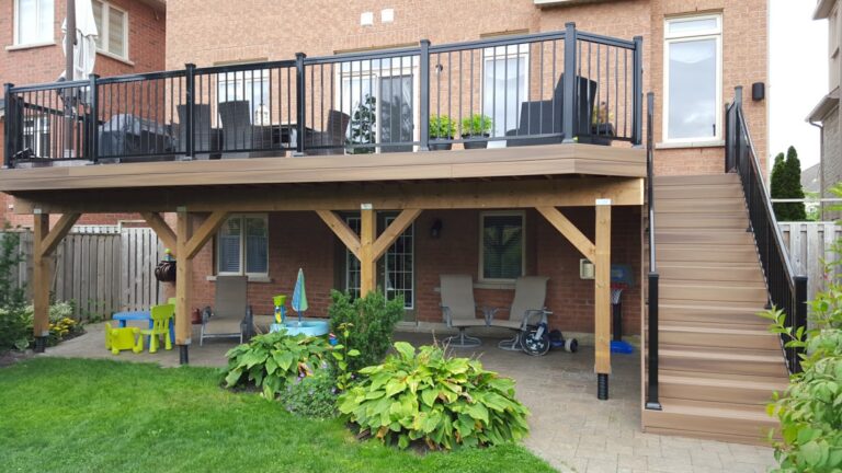 A backyard with a raised wooden deck featuring black PVC railings and stairs. Underneath, there are patio chairs, a small table, and children’s play furniture on a paved area, surrounded by green plants and grass.
