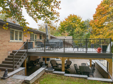 A two-story backyard patio with outdoor seating areas on both levels, black Century Aluminum Pickets railings, stairs, and a red watering can. Trees with autumn leaves surround the space.