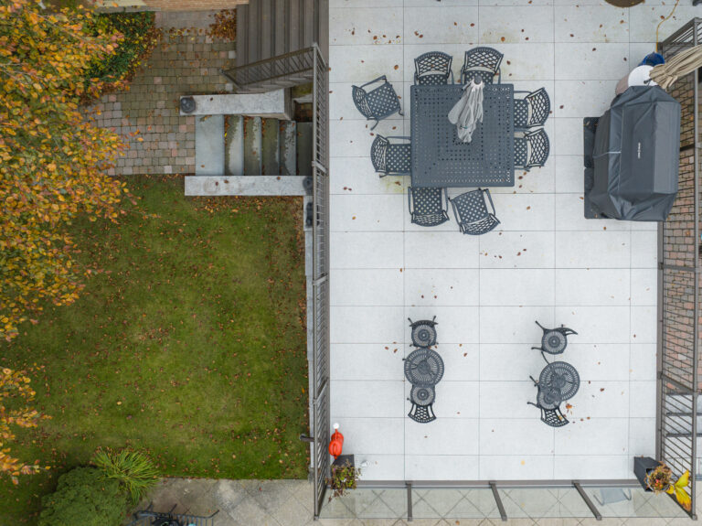 Aerial view of a patio with black metal tables and chairs, an umbrella, a covered grill, and a tiled floor. Adjacent is a grassy area with trees and fallen leaves, separated by stairs and a stone walkway.