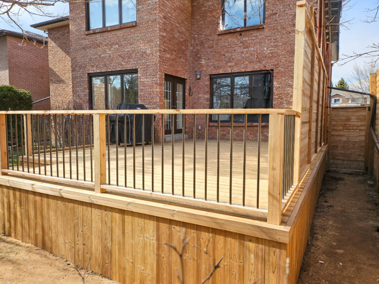 A newly built wooden backyard deck with vertical metal balusters, attached to a brick house with large windows and a patio door. The deck is raised and has a privacy screen on one side.