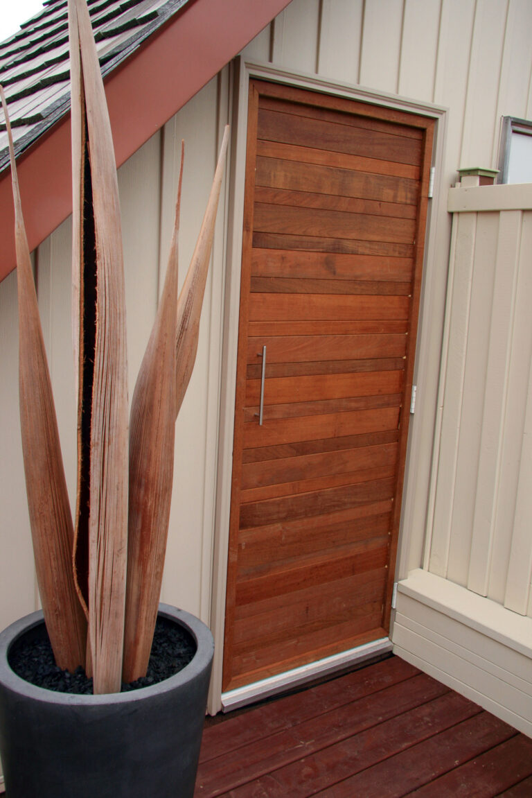 A modern wooden door with a metal handle is set in a beige paneled wall beside a large potted plant on a deck.