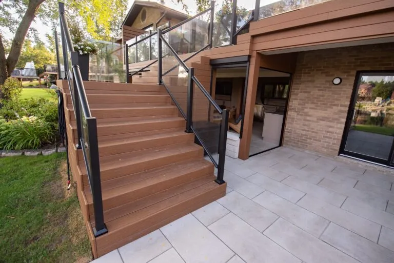 Brown wooden stairs with black railings lead from a glass-walled deck to a paved patio next to a brick house.