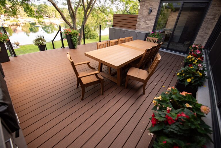 A wooden patio deck with a long dining table, chairs, potted flowers, and a view of a lake in the background.