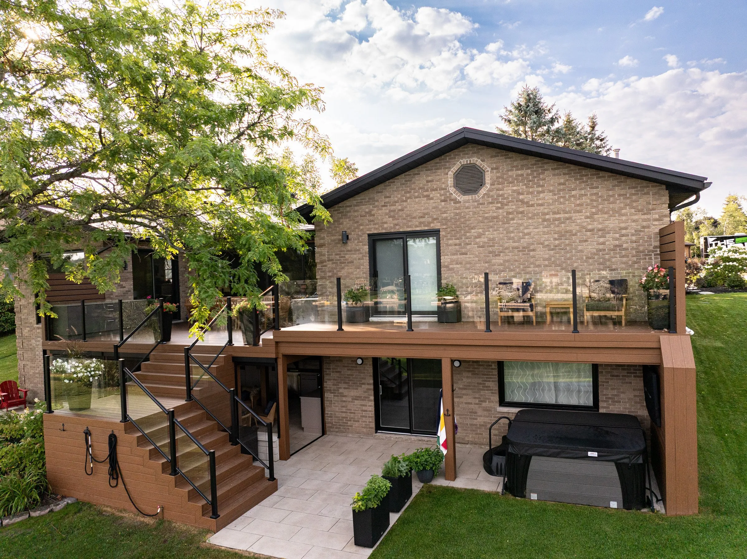 Two-story brick house with glass balcony, patio, stairs, outdoor hot tub, and landscaped yard on a sunny day.