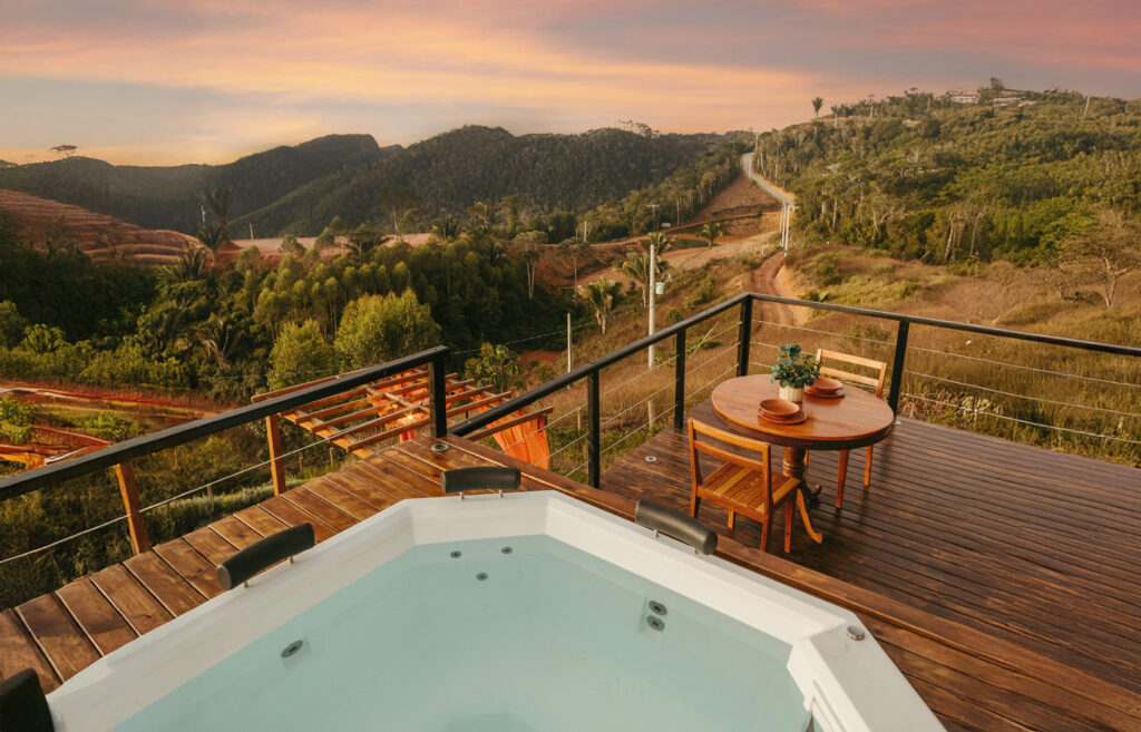 A wooden deck overlooking a scenic landscape features a hot tub and a small round table with chairs. The view includes rolling hills and a vibrant sunset sky in the background, offering a peaceful and relaxing atmosphere.